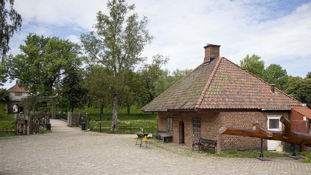 Utsikt mot Glasshytta i Gamlebyen Fredrikstad, omgitt av trær og brosteinsgate.