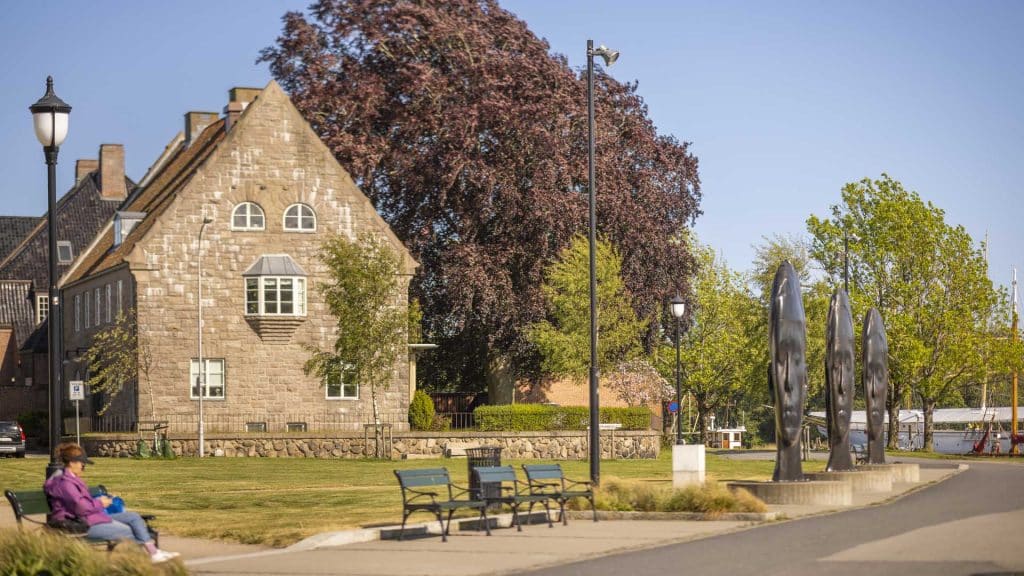 Tre hodeformede skulpturer i mørk bronse står på rekke i en park ved Fredrikstad bibliotek