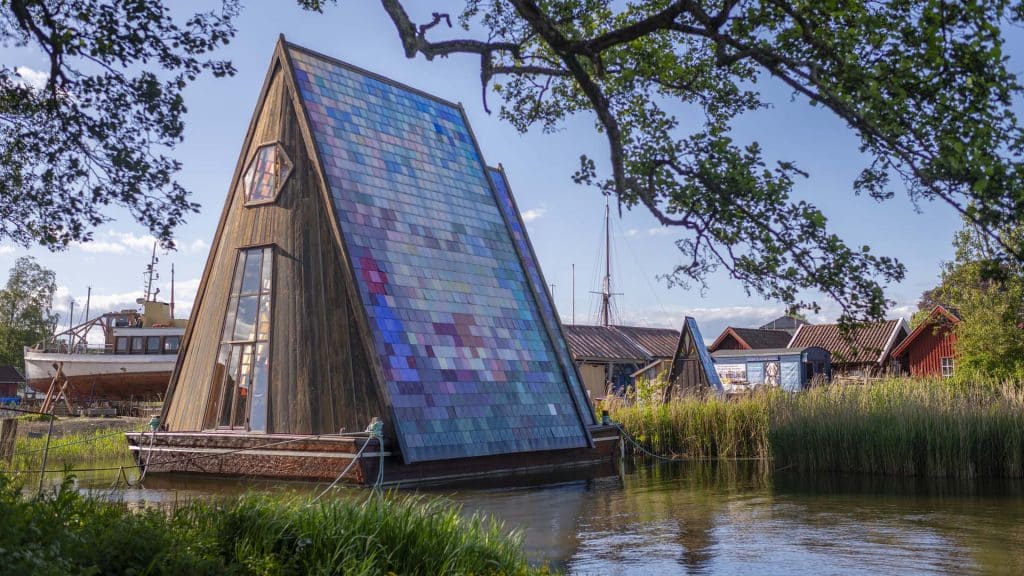 Håpets katedral på Isegran, et trekantformet bygg med fargerike plastfliser, ligger idyllisk til ved vannkanten.