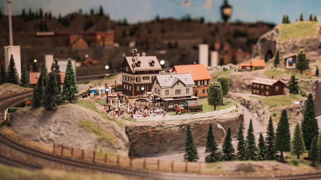 Modelljernbaneanlegg med fjell, trær, jernbanelinjer og små hus, med et lite torg fullt av figurer i aktivitet.