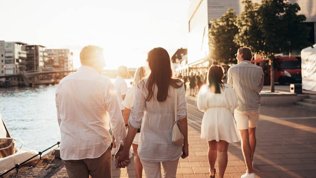 En varm sommerkveld ved brygga i Fredrikstad, der folk går langs elvepromenaden i kveldssol. Bildet viser den livlige stemningen ved vannkanten, omgitt av restauranter, båtliv og moderne bymiljø.