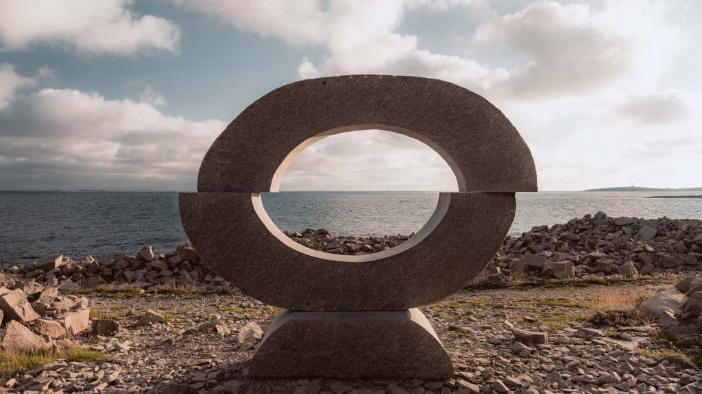 Granittskulptur med sirkelåpning plassert ved kysten på Hvaler, med havet i bakgrunnen.