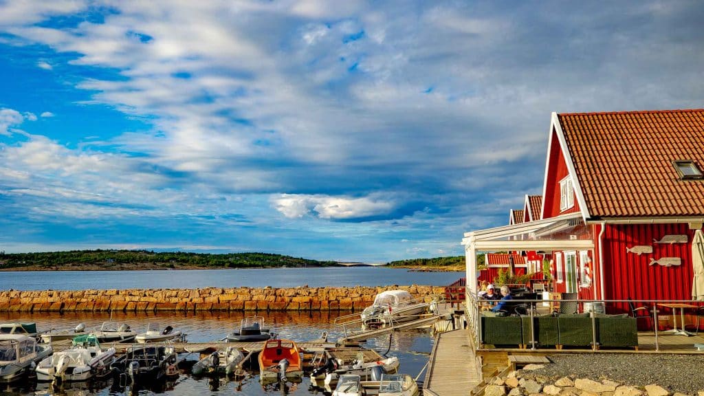 Uteservering ved sjøen og båthavn med rødmalte bygg og blå himmel.