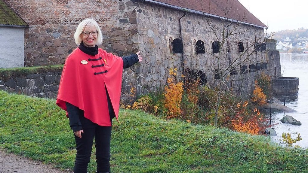 Guide i rød kappe peker mot festningsmur i Gamlebyen, Fredrikstad