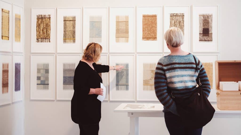 Utstilling med tekstilkunst der en formidler viser detaljer til besøkende.