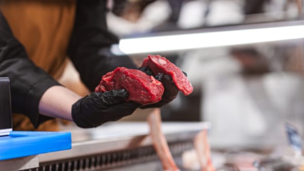 Faglært kjøttansatt holder frem to stykker mørt rødt kjøtt bak disken hos Manstad Kjøtt