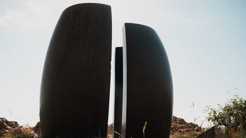 Mørk skulptur i to deler står i et åpent steinlandskap omgitt av vrakstein og natur.