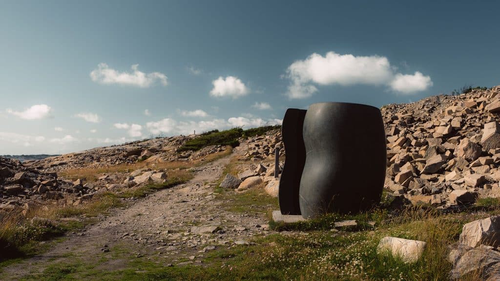 Mørk skulptur i to deler står i et åpent steinlandskap omgitt av vrakstein og natur.