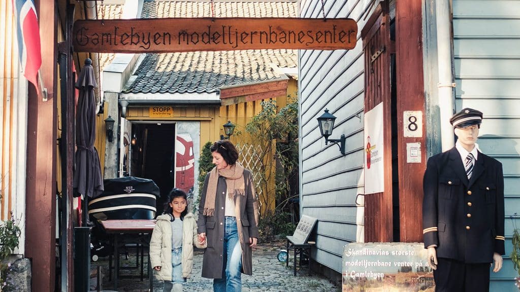 Inngangspartiet til Gamlebyen modelljernbanesenter, med en voksen og et barn som holder hender og en togkonduktør-dukke ved døra.