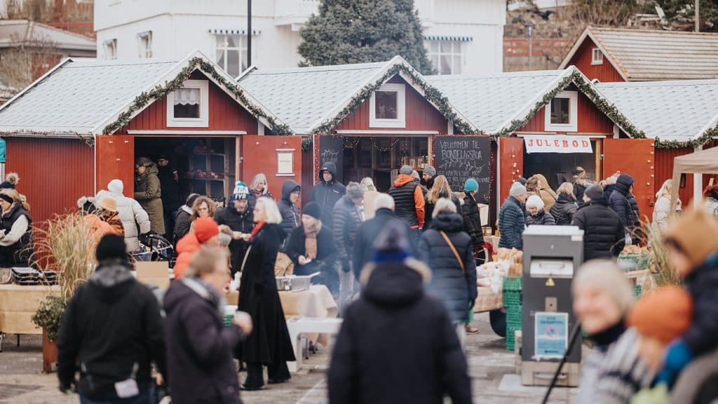 Oversiktsbilde av flere juleboder pyntet med granbar og lys, omgitt av mange besøkende.
