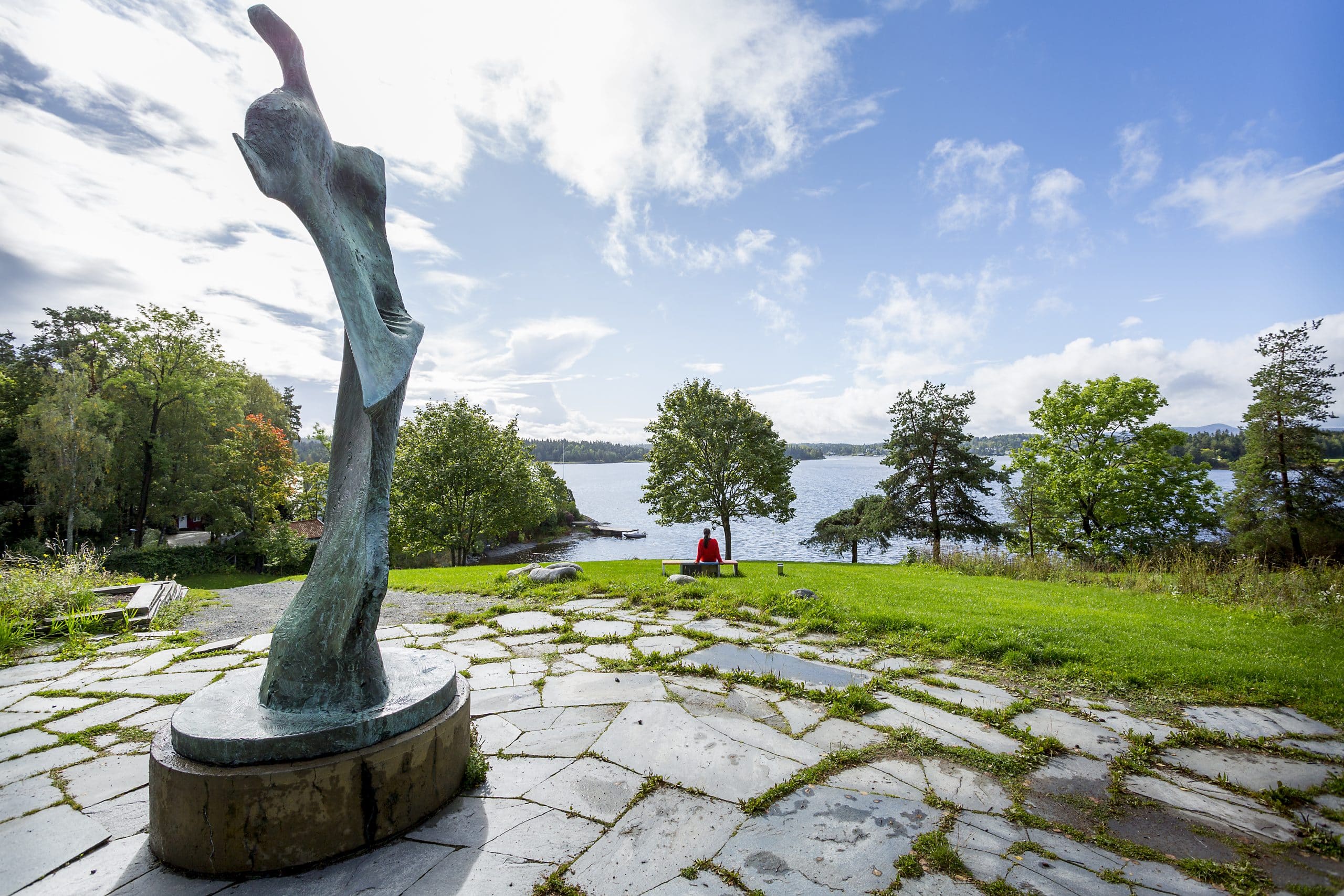 Person i rødt sitter på benk med utsikt mot Oslofjorden, omgitt av gressplen, trær og skulptur i forgrunnen ved Henie Onstad Kunstsenter.