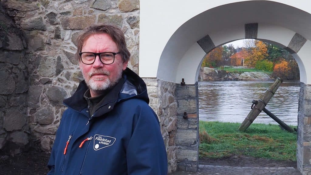 Guide Arild står ved en Kongeporten i Gamlebyen med elven og høsttrær i bakgrunnen.