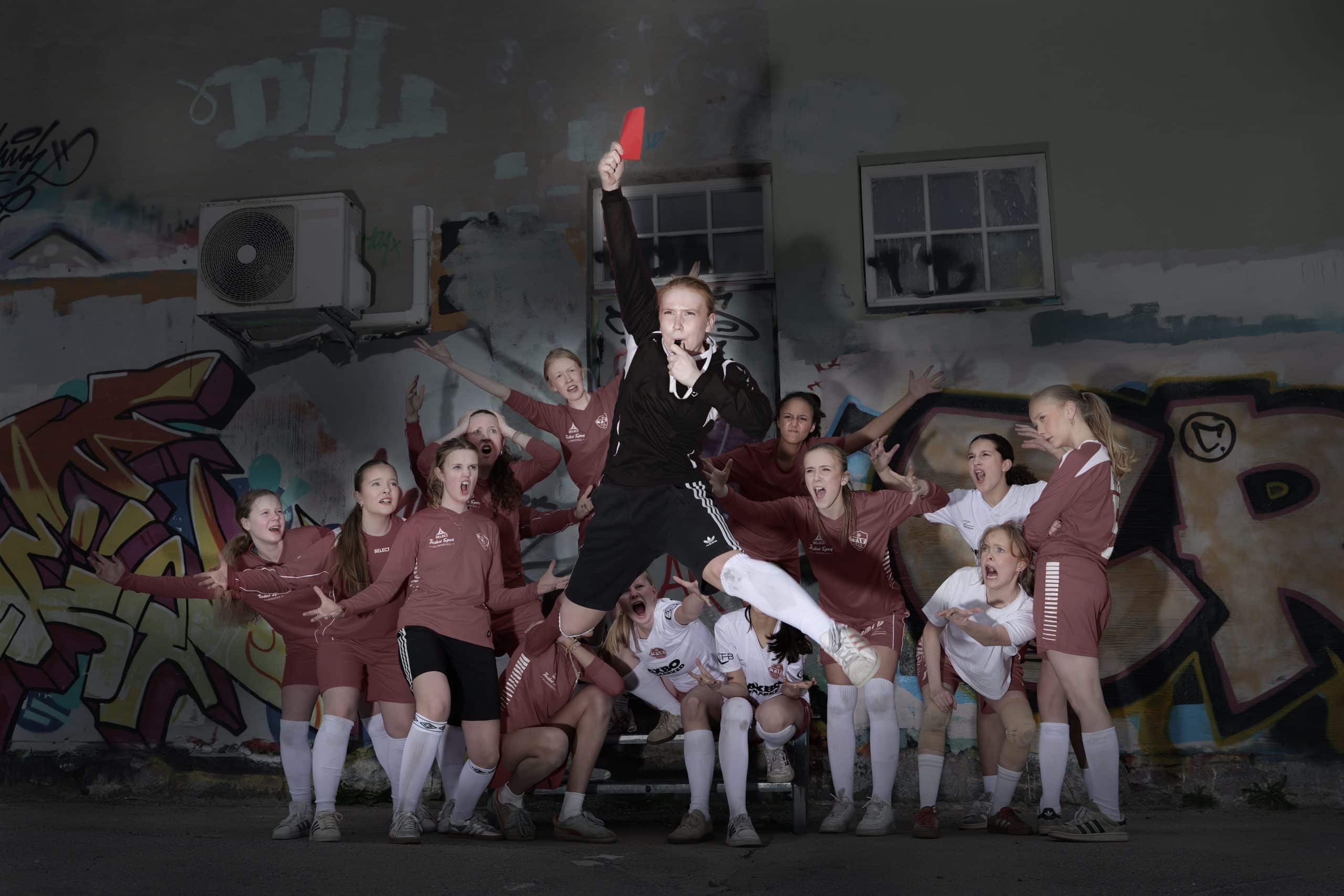 Ungdommer i fotballdrakter foran en graffitivegg. En spiller hopper i lufta mens en dommer viser rødt kort, omgitt av resten av laget.